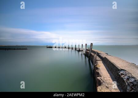 Verwitterte konkrete Wellenbrecher mit einer rostigen Spundwände und schräger Holzbalken im Spiegel glatten Wasseroberfläche auf einer windstillen Tag mit einem bl wider Stockfoto