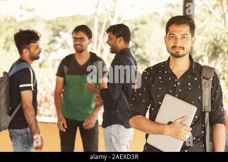 Lächelnden jungen erwachsenen Kursteilnehmer mit Laptop in der Hand, während Freunde staning im Hintergrund - Concfident graduate Scholar an der Universität lächelnd Stockfoto