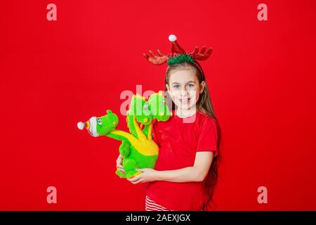 Porträt eines lustigen fröhliches Mädchen mit einem Verband der Hörner auf dem Kopf umarmt einen Plüsch Drache in Weihnachten Schlafanzug isoliert auf einem roten Hintergrund. Stockfoto