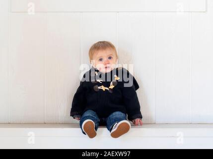 Blonde, blauäugige, Kleinkind Junge auf eine Bank in warme Winterkleidung mit einem natürlichen weißen Holz Hintergrund gekleidet Stockfoto