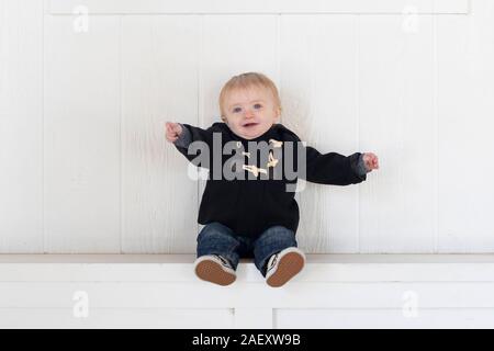 Blonde, blauäugige, Kleinkind Junge auf eine Bank in warme Winterkleidung mit einem natürlichen weißen Holz Hintergrund gekleidet Stockfoto