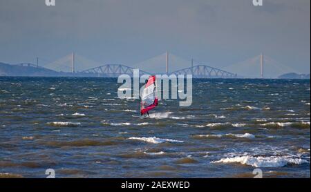 Longniddry Bents, East Lothian, Schottland, Großbritannien. 11. Dezember 2019. Einsame windsurfer Mikey braves 5 Grad Real Feel-3 Temperatur und Wind bei 39 km mit Böen von 58 km/h/h SW seinen Sport im Sonnenschein auf Forth Estuary mit der Burg und der Stadt Edinburgh und Forth Rail Bridge und South Queensferry Kreuzung im Hintergrund zu genießen. Stockfoto