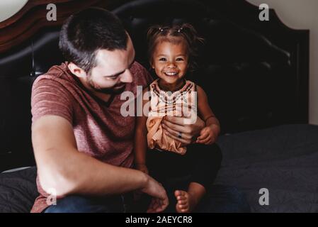 Schöne 2-jährigen gemischtrassigen Mädchen sitzen auf dem Bett mit Dad lächelnd Stockfoto