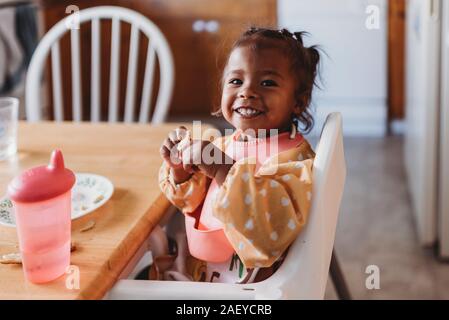 Lächelnd 2 Jahre alten biracial Mädchen mit Bib im Hochstuhl in der Küche Stockfoto