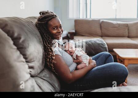 Mitte-30 â € ™ s ethnischen Mama mit langen Zöpfen auf der Couch Holding multirassischen Baby Stockfoto