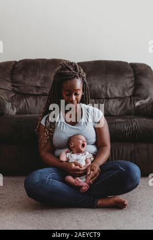 Junge ethnische Mom mit Zöpfen auf dem Boden Holding gähnende Kind Stockfoto