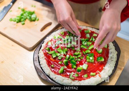 Backblech mit rohen Teig Teig vor dem Backen mit glutenfreien Mehl und Frau Hände gehackte Hinzufügen grüne Paprika in Tomatensauce gemacht Stockfoto