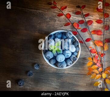 Gefrorene Heidelbeeren in einer weissen Schüssel mit Blätter im Herbst wie kostenlose Farben auf einem strukturierten hölzernen Tisch. Stockfoto