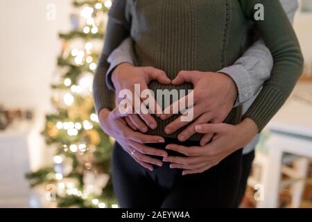 Schwangere Frau und ihr Mann umarmte ihre Bauch mit Weihnachtsbaum im Hintergrund. Stockfoto