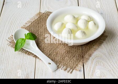 Glasschale mit kleinen Mozzarella Kugeln in einer Salzlake und Porzellan Löffel auf einem weißen rustikalen Holztisch. Healhy mediterrane Küche. Stockfoto