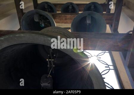 Antient Bronzeglocken hängen auf Holzbalken und Klingeln. Strahlende Sonne und Sonnenstrahlen auf der linken Seite des Bildes. Grau, Braun und Blau. Stockfoto