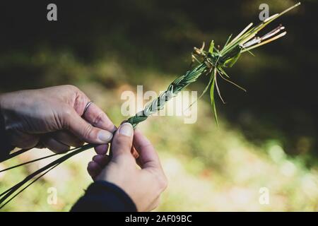 Nahaufnahme der Kaukasischen frau Hände stricken ein grünes Gras traditionelle Diadem Kranz mit Knoten Stockfoto