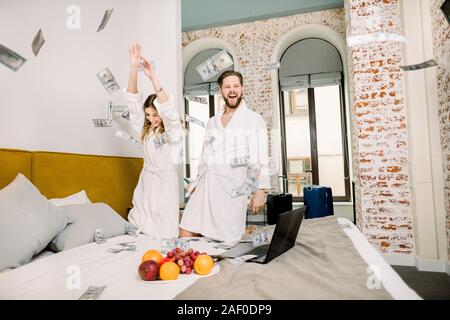 Sehr freundliches junges Paar mit Laptop auf dem Bett und Werfen von Euro oder Dollar Geld in die Luft. Glückliches Paar mit dem fallenden Dollar Banknoten Stockfoto