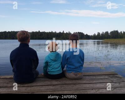 Drei Jungen verschiedenen Alters in blauer Pullover in verschiedenen Farben sitzen auf einem warmen Ponton aus Holz, Beine baumeln, ihren Rücken auf den Betrachter, auf den See und die grünen Wald dahinter suchen. Stockfoto