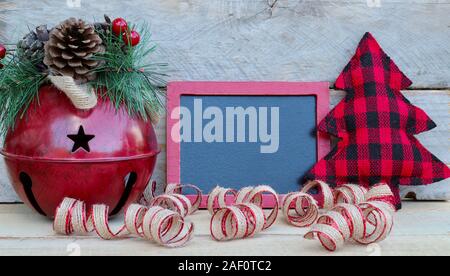 Red Jingle Bell mit Pinien und Beeren neben einem rot eingerahmten schwarzen Brett, Tuch, Weihnachtsbaum und gewelltes Band auf einem Holz Hintergrund mit Kopie Raum Stockfoto