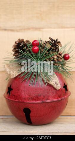 Große rote Jingle Bell dekoriert mit Pine Cone, Beeren und Bastschleife vertikal auf einer natürlichen Holz Hintergrund mit Kopie Raum Stockfoto