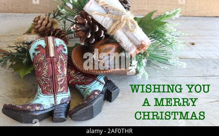 Cowboystiefel und Hut mit einem Pine Cone Spray und gebundene Bündel Holz flach auf einem Holz Hintergrund mit wünschen Ihnen ein frohes Weihnachtsfest in grüner Schrift Stockfoto