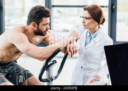 Arzt in der Nähe von Sportsman mit Elektroden im Fitnessraum während Dauertest Stockfoto