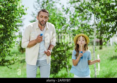 Schöner Mann und Tochter in Strohhut bläst Seifenblasen in der Nähe von Bäumen Stockfoto