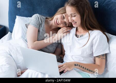 Zwei lächelnde Lesben umarmt, während mit Laptop im Bett am Morgen Stockfoto