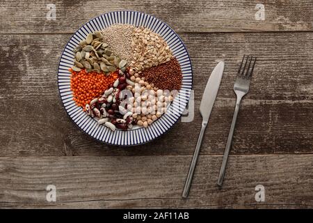 Blick von oben auf die Gabel und Messer in der Nähe von keramikplatte mit rohen sortierten Bohnen, Getreide und Saaten auf dunklen Holz- Oberfläche mit Kopie Raum gestreift Stockfoto
