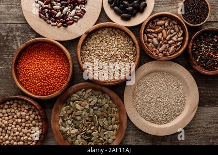 Blick von oben auf die Schüsseln mit rohen Haferflocken, rote Linsen, verschiedene Bohnen, Quinoa, Kichererbsen, Pfefferkörner und Kürbiskerne auf Holz- Oberfläche Stockfoto