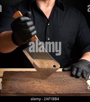 Koch in einem schwarzen Hemd und schwarzen Latex Handschuhe hält eine große Küche Messer zum Schneiden von Fleisch, Low Key Stockfoto