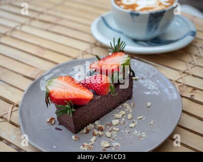 Köstliche Schokolade Torte mit Erdbeeren und Beeren auf Graue Keramik Schüssel mit heißem Kaffee Tasse auf Bambus Tisch lagen. Stockfoto