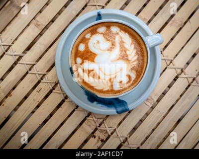 Heißer Kaffee Tasse mit Latte Art Kaffee mit Zirkus Affe sitzt auf Einrad Kugeln und spielen mit Blättern, latte Art in blau Keramik Schale auf Bambus ba Stockfoto