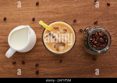 Blick von oben auf Eis Kaffee mit Stroh in der Nähe von Milch und Kaffee in der Kanne Körner in Glas auf hölzernen Tisch Stockfoto