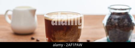 Selektiver Fokus der Eis Kaffee mit Milch in Glas in der Nähe von Milchkännchen und Kaffee Körner auf Holztisch auf Weiß, Panorama-aufnahme isoliert Stockfoto