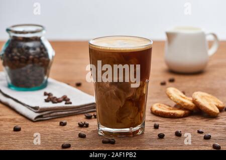 Selektiver Fokus der Eis Kaffee mischen mit Milch in Glas in der Nähe von milchkännchen, Plätzchen und Kaffee Körner auf Holztisch isoliert auf Grau Stockfoto