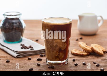 Selektiver Fokus der Eis Kaffee mischen mit Milch in Glas in der Nähe von milchkännchen, Plätzchen und Kaffee Körner auf Holztisch isoliert auf weißem Stockfoto