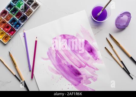 Blick von oben auf die Lila aquarell Pinselstrichen auf weißem Papier in der Nähe von Pinseln, Farben und Buntstifte auf Marmor weiß Oberfläche Stockfoto