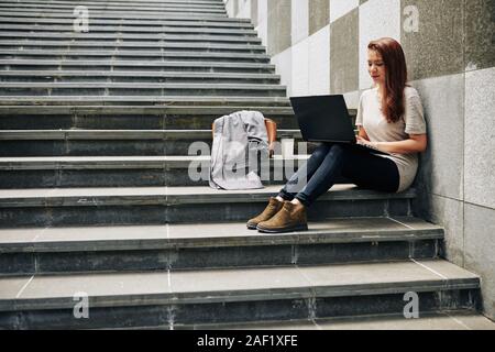Lächelnd weibliche Freiberufler sitzen auf Schritte im Freien, Arbeiten am Laptop und das Schreiben von Artikeln Stockfoto