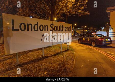 Beleuchtetes Schild am Flughafen London Southend bei Nacht mit Straßenlaternen, Baum und vorbeifahrendem Taxi in der Nähe der Zufahrtsstraße in Essex, Großbritannien. Stockfoto