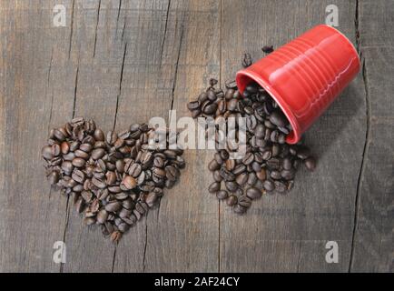 Stapel von Kaffeebohnen bilden ein Herz neben einem Verschüttete rote Schale auf rustikalen Holzmöbeln Hintergrund Stockfoto