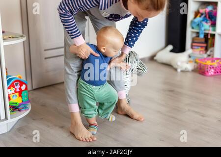 Süße kleine adorable Blonde toddler Boy machen erste Schritte mit Mutter Unterstützung im Spielzimmer zu Hause. Gerne lustig Kind lernen mit Mama helfen zu gehen Stockfoto