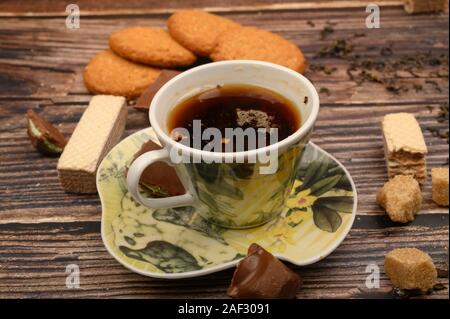 Eine Tasse schwarzer Tee, Teeblätter, Stücke von braunem Zucker und Haferflocken Kekse, Waffeln, Stücke Schokolade auf einem hölzernen Hintergrund. Nahaufnahme Stockfoto