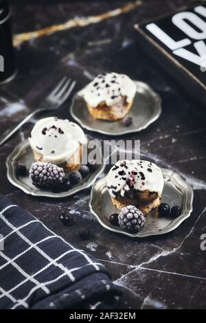 Leckere blueberry muffins mit weißer Schokolade Sahnehäubchen auf Zinn Tabletts mit Beeren auf schwarzem Marmor Hintergrund eingerichtet Stockfoto