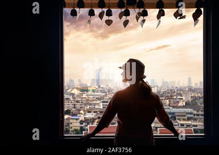 Silhouette der Frau in der Hut ist auf der Suche nach Bangkok im Fenster Blick auf die Stadt der Wolkenkratzer Geschäftsviertel von Goldenen Berg Pagoda Wat Saket bei Orange Stockfoto