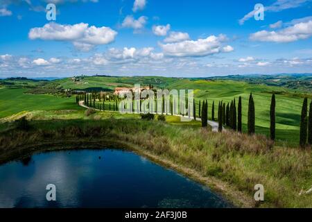 Eine gewundene Schotterpiste, flankiert von Cypress Alleen, vorbei an einem kleinen See, der zum Agriturismo Baccoleno Stockfoto