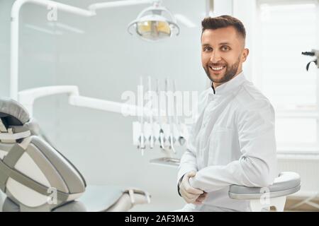 Seitenansicht der professionellen bärtigen Zahnarzt in weiße Uniform sitzen in Zahnarztpraxis und posieren. Mann mit Kamera und lächelnd, während in kieferorthopädischen Klinik arbeiten. Konzept der Medizin und Job. Stockfoto