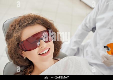 Blick von oben der schönen Frau liegend auf zahnmedizinischen Stuhl in eine Schutzbrille. Curly Patientin lächelnd, während sie die Verfahren. Konzept der Zahnmedizin und die Zähne. Stockfoto