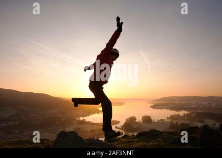 Ein Mann springen vor Freude bei Sonnenaufgang über Lake Windermere von Todd Crag über Ambleside, Lake District, Großbritannien. Stockfoto