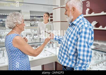 Ältere Dame mit grauen Haaren trägt blaue Polka Dot Dress, die versuchen, auf Perle Halskette und Spiegel, dass ihr Mann mit kariertem Hemd. Gerne reifes Paar kaufen Schmuck in Luxus store Stockfoto