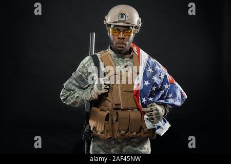 Vorderansicht des tapferen afrikanische Soldaten mit National Flagge auf der Schulter und Waffe posiert. Ernst ranker tragen Uniform, Helm und Brille mit Kamera schaut. Konzept der Armee. Stockfoto