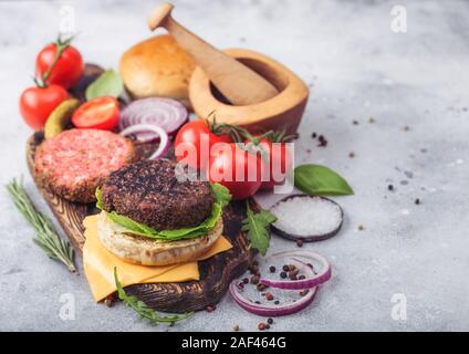 Frisches rohes Hackfleisch und gegrillte Paprika Rindfleisch Burger auf vintage Schneidbrett mit Brötchen und Zwiebeln und Tomaten auf Holzuntergrund. Mörser mit Pistill mit p Stockfoto