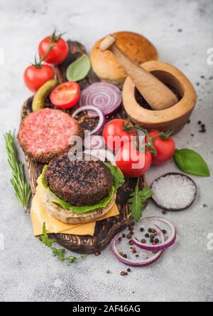 Frisches rohes Hackfleisch und gegrillte Paprika Rindfleisch Burger auf vintage Schneidbrett mit Brötchen und Zwiebeln und Tomaten auf Holzuntergrund. Mörser mit Pistill mit p Stockfoto