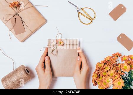 Weibliche Hände halten hausgemachte Kraftpapier Geschenk Tasche neben dem Feld, Jute Seil und Strauß Blumen auf weißem Tabelle, Ansicht von oben. Stockfoto